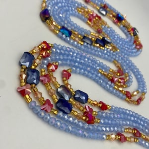 May include: Pale blue beaded necklaces with gold-coloured accents and colourful gemstone details. The necklaces feature strands of small, faceted beads, interspersed with larger, geometric blue and red beads.