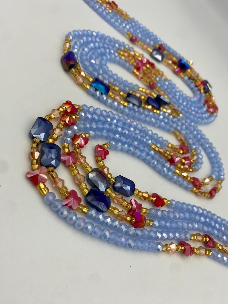 May include: Pale blue beaded necklaces with gold-coloured accents and colourful gemstone details. The necklaces feature strands of small, faceted beads, interspersed with larger, geometric blue and red beads.