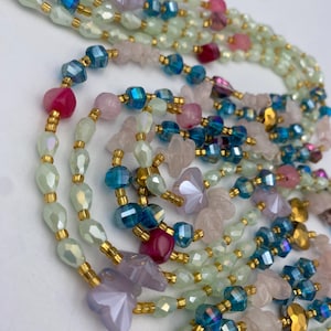 May include: Close-up of layered beaded necklaces in various colors. The necklaces feature iridescent green, pink, blue, and gold beads. Some beads are faceted, and others are shaped like flowers. The necklaces are strung on gold-colored wire.