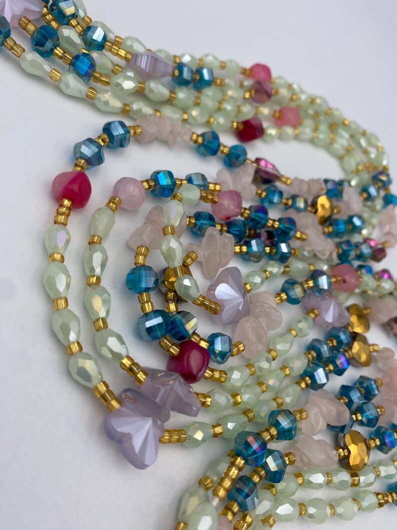 May include: Close-up of layered beaded necklaces in various colors. The necklaces feature iridescent green, pink, blue, and gold beads. Some beads are faceted, and others are shaped like flowers. The necklaces are strung on gold-colored wire.