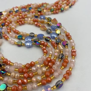 May include: Close-up of several strands of beaded jewellery. The beads are in shades of peach, pink, blue, and iridescent colours. The beads are strung on gold-coloured wire, creating a delicate and elegant look.