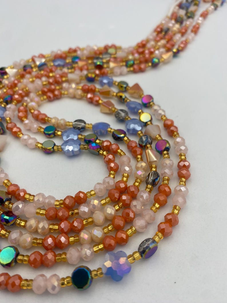 May include: Close-up of several strands of beaded jewellery. The beads are in shades of peach, pink, blue, and iridescent colours. The beads are strung on gold-coloured wire, creating a delicate and elegant look.