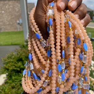 May include: A multi-strand beaded necklace with peach-colored beads, blue beads, and clear glass beads. The necklace is made of multiple strands of beads and is held in a hand.