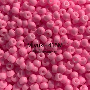 May include: A close-up image of a large quantity of pink seed beads. The beads are small and round, with a hole in the center. The beads are arranged in a random pattern, creating a textured surface. The text "Miyuki-415M Maddiebaysshop" is visible in the center of the image.