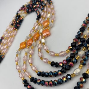 May include: A multi-strand beaded necklace in various colours. The necklace features black, pink, orange, and white beads, with gold-coloured spacers. The beads are arranged in multiple strands, creating a layered effect. The necklace is designed to be worn as a statement piece.