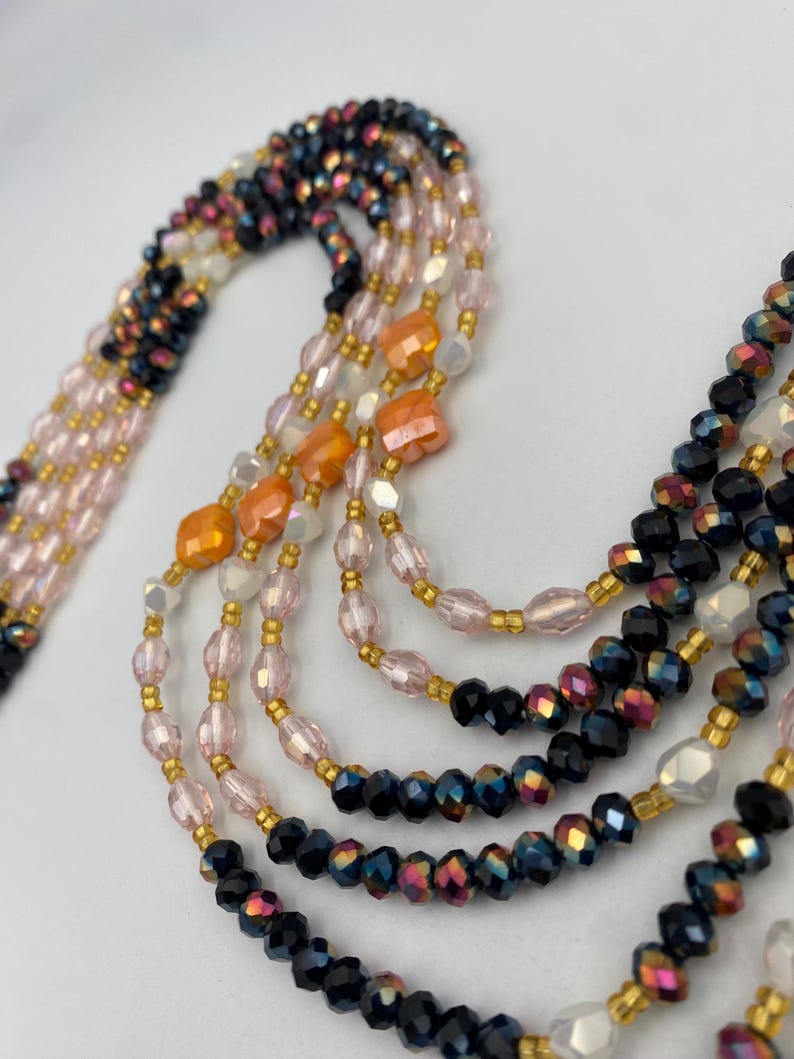 May include: A multi-strand beaded necklace in various colours. The necklace features black, pink, orange, and white beads, with gold-coloured spacers. The beads are arranged in multiple strands, creating a layered effect. The necklace is designed to be worn as a statement piece.