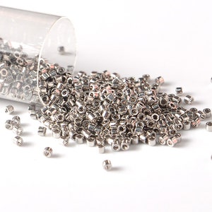 May include: A large quantity of silver-colored seed beads are scattered on a white surface. The beads are small and round, and they are spilling out of a clear glass container.