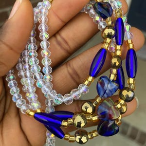 May include: A selection of waist beads in a hand. The beads are clear, iridescent, gold, and deep blue. Some beads are heart-shaped. The waist beads are strung together on a clear string.