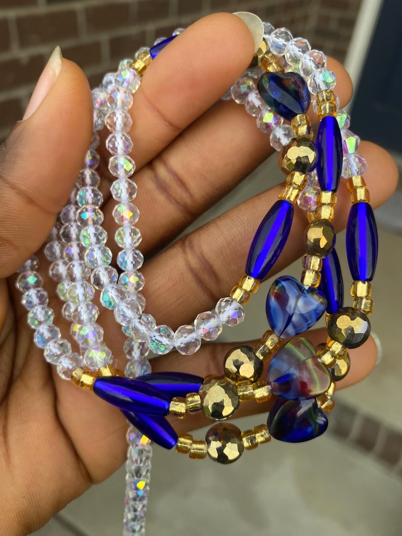 May include: A selection of waist beads in a hand. The beads are clear, iridescent, gold, and deep blue. Some beads are heart-shaped. The waist beads are strung together on a clear string.