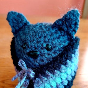 May include: A crocheted stuffed animal in the shape of a wolf wearing a blue and purple scarf. The wolf has black eyes and a black nose.