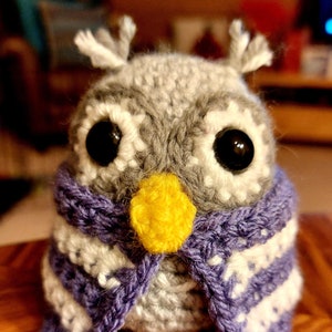 May include: A gray crocheted owl wearing a purple and white striped scarf. The owl has black eyes and a yellow beak.