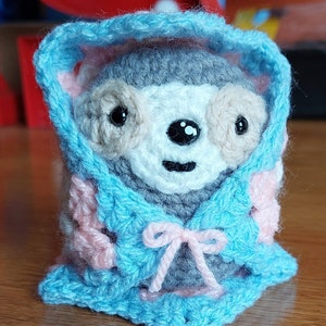 Peut inclure: Un animal en peluche crocheté portant une veste à capuche bleue avec des accents roses. L'animal a un visage gris avec de la fourrure blanche et des yeux noirs.