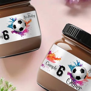 Op de afbeelding: Twee glazen potten gevuld met een bruine substantie, elk met een zwart deksel. De potten hebben labels met een voetbalafbeelding, kleurrijke verfspatten en de tekst "Happy Birthday" en "Is turning 6".
