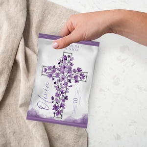 May include: A white and purple favor bag with a cross design. The cross is decorated with purple flowers and vines. The bag has the name "Olivia" in cursive and the text "PRIMERA COMUNION".