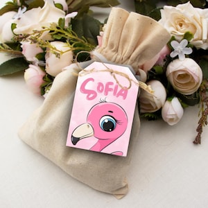 May include: A beige drawstring bag with a pink tag featuring a cartoon flamingo and the name "SOFIA" in pink lettering. The tag is tied with twine. The background includes white and pink roses.