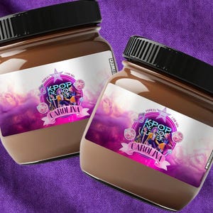 Op de afbeelding: Twee potten chocoladekleurige spread met zwarte deksels. Elke pot heeft een etiket met de tekst "KPOP DE CAROLINA" in roze en wit, met een afbeelding van mensen en muzieknoten. De achtergrond is een paars en roze ontwerp.