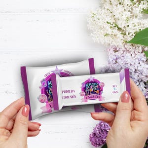 May include: Two white and purple candy bar wrappers with text and graphics. One wrapper is in the background, and the other is held in a person's hand. The wrappers have the text "KPOP de Gloria" and "Primera Comunión" with a star and musical note design.