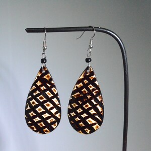 Puede incluir: Un par de pendientes en forma de lágrima con un patrón geométrico negro y marrón. Los pendientes están hechos de madera y tienen ganchos plateados.