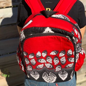 Puede incluir: Una mochila roja con tejido estampado en blanco y negro. La mochila tiene un compartimento principal grande con cierre de cremallera y dos bolsillos más pequeños en la parte delantera.