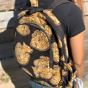 Puede incluir: Mochila negra con un patrón de peces dorados y negros. La mochila tiene un bolsillo con cremallera en la parte delantera y correas ajustables.