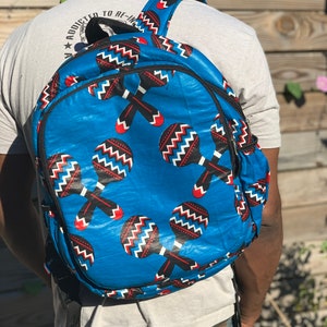 Puede incluir: Una mochila azul con un patrón de maracas. Las maracas son rojas, negras y blancas. La mochila tiene correas negras y un bolsillo con cremallera.