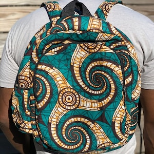Puede incluir: Una mochila con estampado de color verde azulado, marrón y dorado con una cremallera negra. El estampado presenta diseños en espiral.
