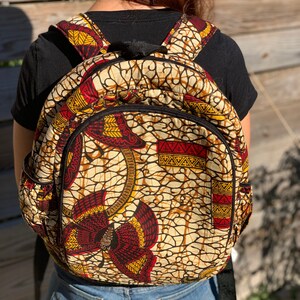 Puede incluir: Una mochila de color beige, rojo y amarillo con una cremallera negra y correas ajustables.