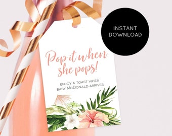 May include: A white gift tag with pink and green tropical floral designs. The tag has the text "Pop it when she pops!" and "Enjoy a toast when baby McDonald arrives".