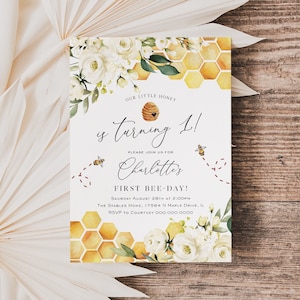 May include: A white and gold birthday invitation with a beehive and bees. The text reads "Our little honey is turning 1! Please join us for Charlotte's First Bee-Day! Saturday August 28th at 2:00pm The Stables Home, 17564 N Maple Drive, IL RSVP to Courtney 000.000.0000"