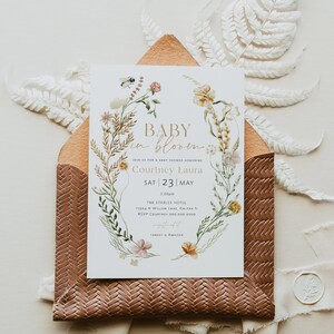 May include: A baby shower invitation featuring a floral wreath design. The invitation reads "Baby in Bloom" and includes the name "Courtney Laura" and the date "Sat 23 May".