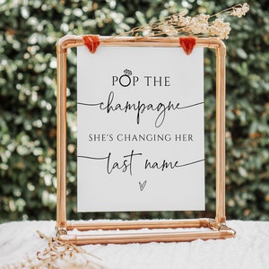 Könnte beinhalten: Ein weißes Schild mit schwarzem Text, der "Pop the champagne she's changing her last name" lautet, mit einem kleinen Diamantenring-Symbol über dem Wort "Pop". Das Schild wird auf einem Kupferrohrständer angezeigt.