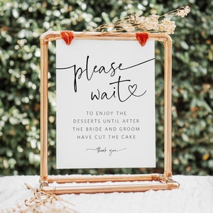May include: A white sign with black text that reads "please wait" with a heart symbol. The sign is on a copper pipe stand and says "To enjoy the desserts until after the bride and groom have cut the cake. Thank you."