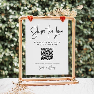 以下が含まれることがあります： 「Share the Love」と「Please share your photos with us」という黒い文字が書かれた白い看板。テキストの下には白黒のQRコードがあります。看板は銅製のスタンドの上に置かれています。看板の下部には「Scan with your smartphone and upload」と「Sarah + Anthony」というテキストが書かれています。