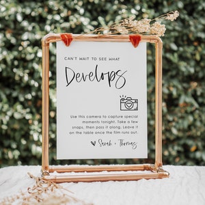 Puede incluir: Un cartel blanco con un marco de cobre y una cinta de terciopelo rojo cuelga de un soporte de tubería de cobre. El cartel dice "Can't wait to see what develops" con un dibujo de una cámara con un corazón dentro. El cartel también dice "Usa esta cámara para capturar momentos especiales esta noche. Toma algunas fotos, luego pásala. Déjala sobre la mesa una vez que la película se acabe. Sarah Thomas"
