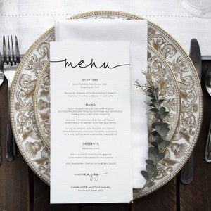 May include: A white menu card with black text on a table setting with two gold and white patterned plates, silverware, and a sprig of greenery. The menu lists "Starters", "Mains", "Desserts", and "Enjoy" with the names Charlotte and Nathaniel and the date November 28th, 2021.