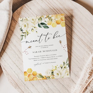 May include: A white card with a watercolour design of white flowers and a honeycomb pattern. The text on the card reads "meant to bee" and includes details for a bridal shower for Sarah McDonald.
