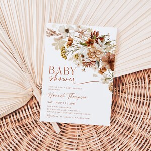 May include: A white baby shower invitation with a floral design in shades of brown and beige. The text reads "BABY SHOWER" in a brown script font. The invitation includes the date, time, and location of the shower, as well as the name of the honoree, Hannah Thompson.