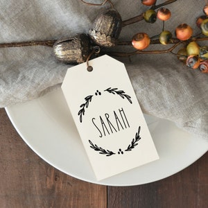 Puede incluir: Una etiqueta blanca con un diseño de corona negra y el nombre "SARAH" impreso en su interior. La etiqueta está colocada en un plato blanco con una servilleta marrón y una rama de bayas.