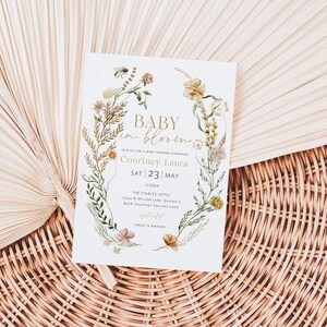 May include: A white baby shower invitation with a floral wreath border. The text reads "Baby in Bloom" and includes the name "Courtney Laura" and the date "Sat 23 | May".