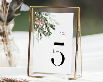 May include: A gold-framed table number card with a white background. The card features a watercolor design of green pine branches, red berries, and the number 5 in a bold black font. The words "TABLE NO." are at the top.