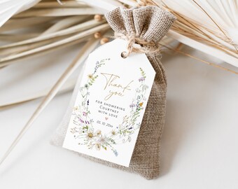 Puede incluir: Una etiqueta blanca de agradecimiento con un diseño de corona floral y letras doradas. La etiqueta dice "Gracias por colmar a Courtney de amor 05.02.20xx". La etiqueta está unida a una pequeña bolsa de arpillera.