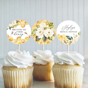 May include: Three cupcakes with white icing, each adorned with a circular decorative pick. The picks display floral and honeycomb patterns, with text reading "She's Found Her Honey" and "Hayley's Bridal Shower". The colour scheme includes yellow, white, and green.