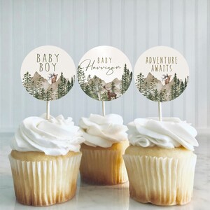 Op de afbeelding: Drie cupcakes versierd met witte frosting en bedekt met ronde papieren cupcake-toppers met een boslandschap met een hert en de tekst "Baby Boy", "Baby Harrison" en "Adventure Awaits".