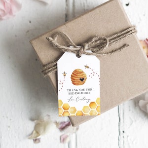 Può includere: Etichetta regalo bianca con un'illustrazione di alveare e il testo "Grazie per essere qui!" con un nome scritto a mano in basso. L'etichetta è legata con uno spago a una scatola regalo marrone.