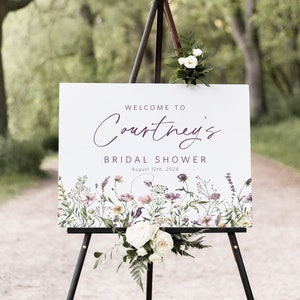 May include: A white sign with purple text that reads "Welcome to Courtney's Bridal Shower August 12th, 2024". The sign is decorated with a watercolor floral border and is displayed on a black easel.
