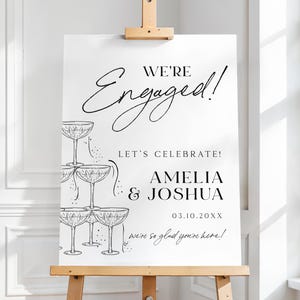 以下が含まれることがあります： 木製のイーゼルに立てられた白い婚約発表のサイン。サインには、エレガントな筆記体で「We're Engaged!」という言葉と、Amelia & Joshuaの名前、日付03.10.20XXが記載されています。シャンパングラスのスタックが描かれています。