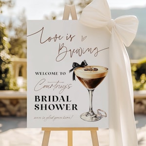 May include: A white sign on a wooden easel with the text "Love is Brewing" and "Welcome to Courtney's Bridal Shower". A cocktail illustration is featured, along with a large white bow and fabric draping.