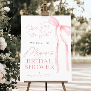 May include: A white bridal shower sign with pink watercolour ribbon and bow. The text reads "She's Tying the Knot!"  "Welcome to Megan's Bridal Shower" and "46 Days until she says 'I Do'."