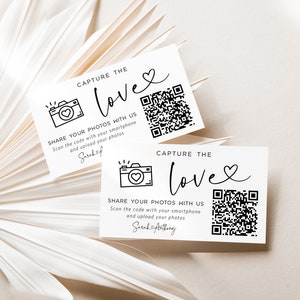 May include: Two white cards with black text and a black and white camera icon. The text on the cards reads "Capture the Love" and "Share your photos with us. Scan the code with your smartphone and upload your photos." The cards also have a QR code.