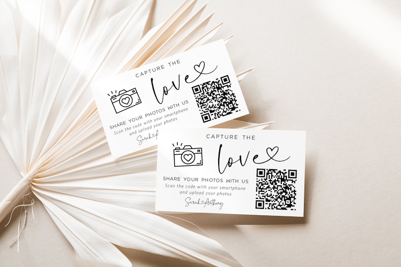 May include: Two white cards with black text and a black and white camera icon. The text on the cards reads "Capture the Love" and "Share your photos with us. Scan the code with your smartphone and upload your photos." The cards also have a QR code.